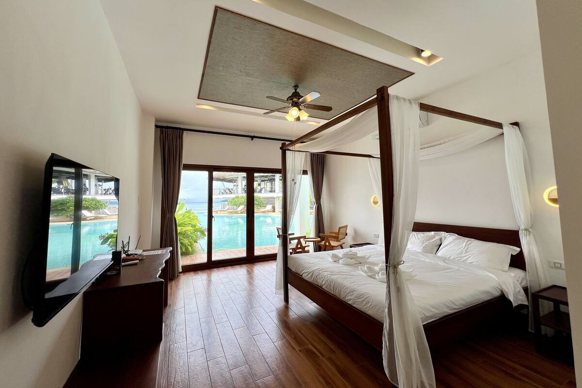 A spacious bedroom with a large bed, a ceiling fan, and a view of a pool and outdoor area.