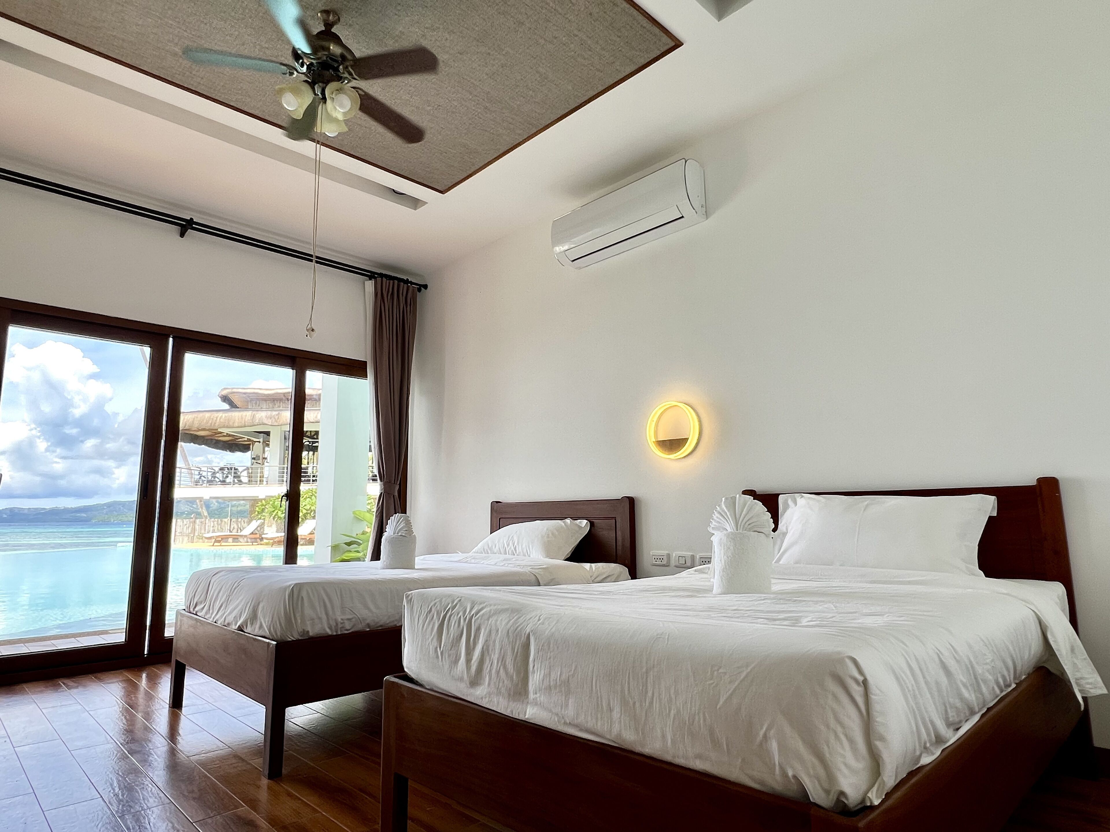 Ein Hotelzimmer mit zwei Betten, einem Deckenventilator, einer Klimaanlage, einem großen Fenster mit Meerblick und einem Balkon mit Meerblick.