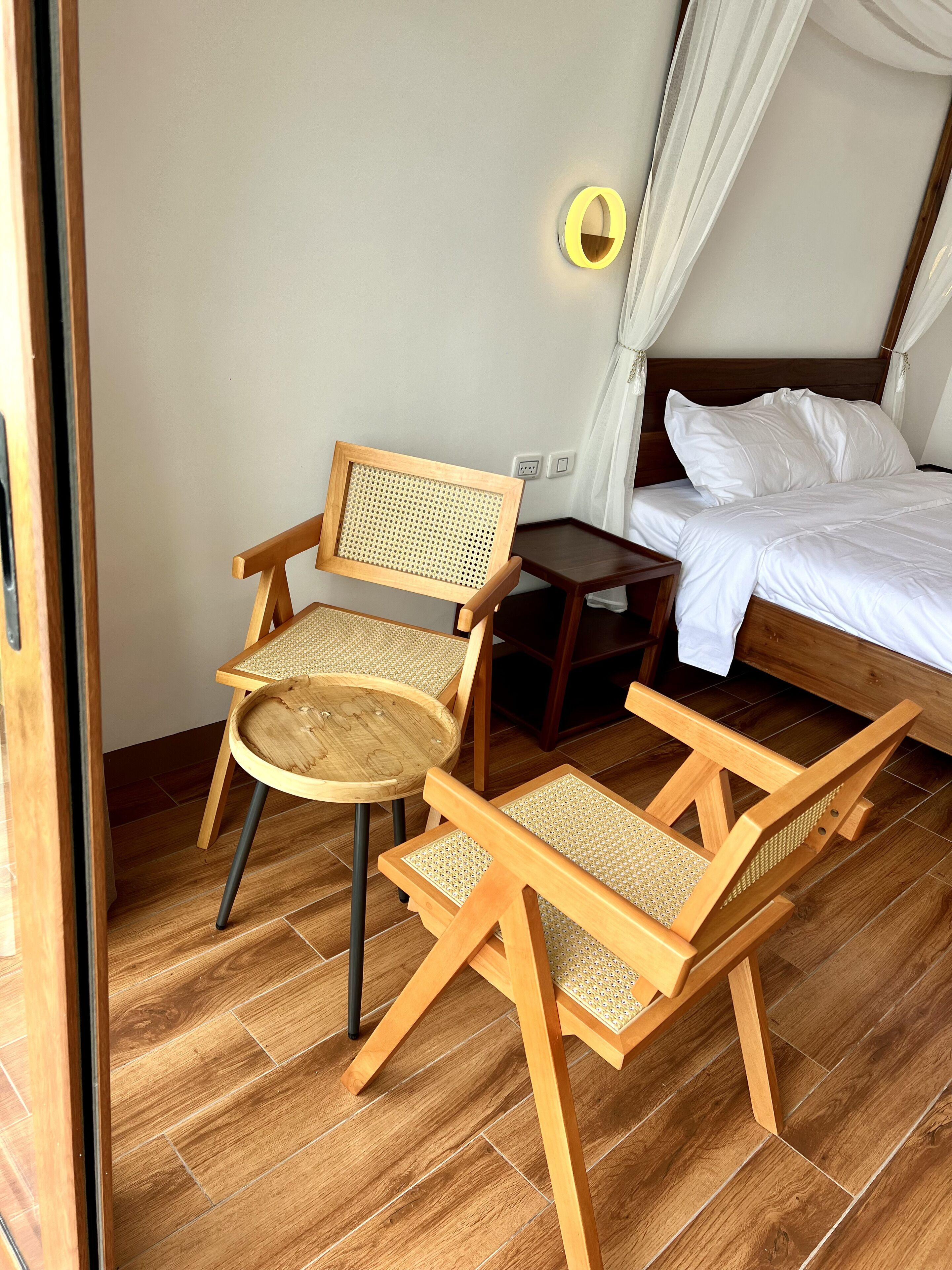 A room with a bed, two wooden chairs with woven seats, a small wooden table, and a wall-mounted light fixture.