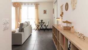 Een woonkamer met een bank, een salontafel en een televisie. Er is een raam met gordijnen, een wandlamp en een kleine plant op de tafel.