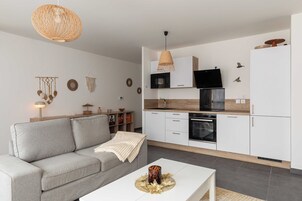 Una sala de estar moderna con un sofá gris, una mesa de centro blanca y una cocina con armarios blancos, un microondas y una estufa.