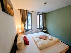 A neatly made bed with pillows, a chair, a window with curtains, and a radiator.