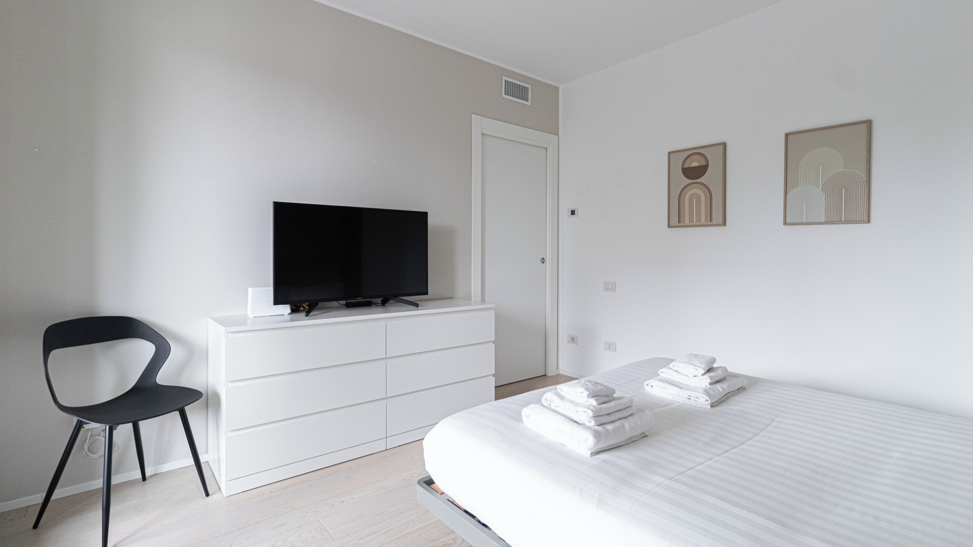A modern bedroom with a bed, a TV on a dresser, a chair, and artwork on the wall.