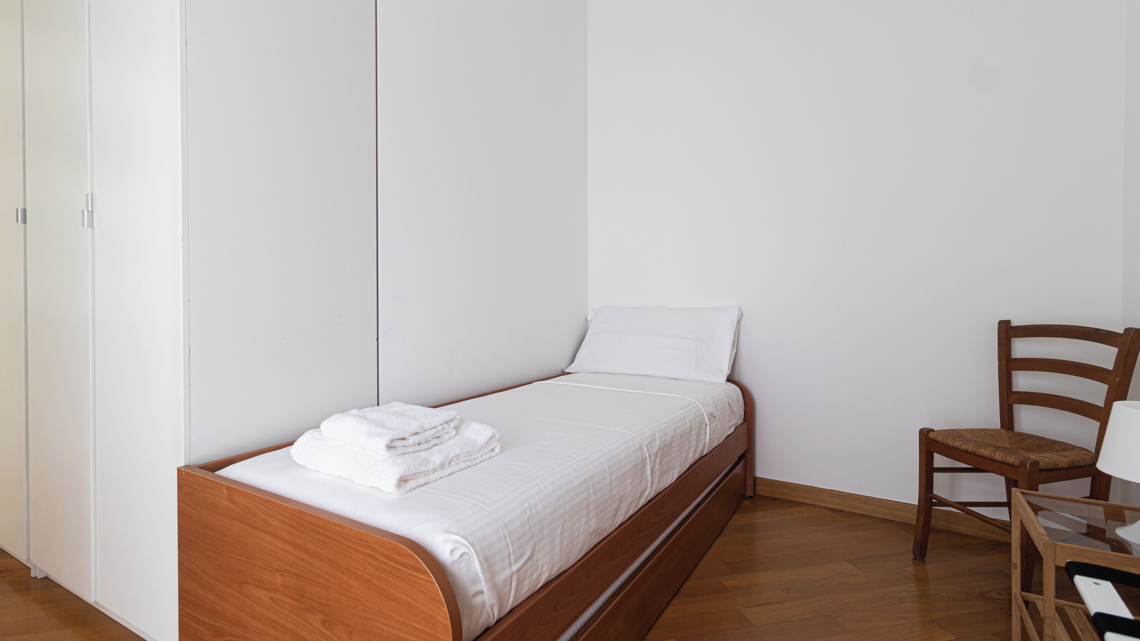 A single bed with a wooden frame, white bedding, and a folded white towel on the footboard. A wooden chair is positioned to the right of the bed.
