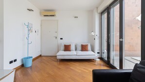 Una habitación de hotel moderna con piso de madera, un sofá blanco con cojines marrones, un sillón de cuero negro y un amplio ventanal con vistas al exterior.