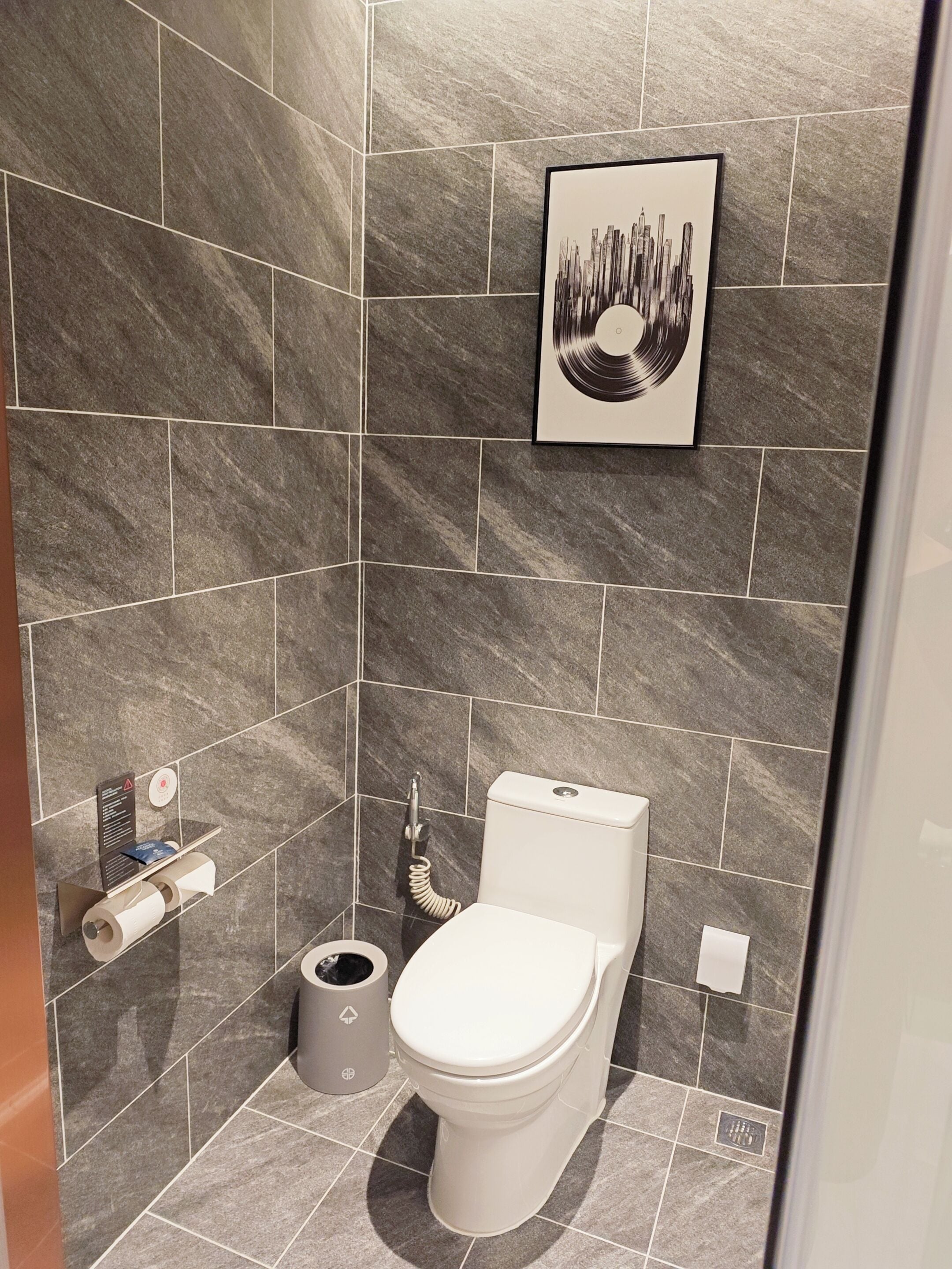 A modern bathroom with grey tiles, a toilet, a hand dryer, and a trash bin.