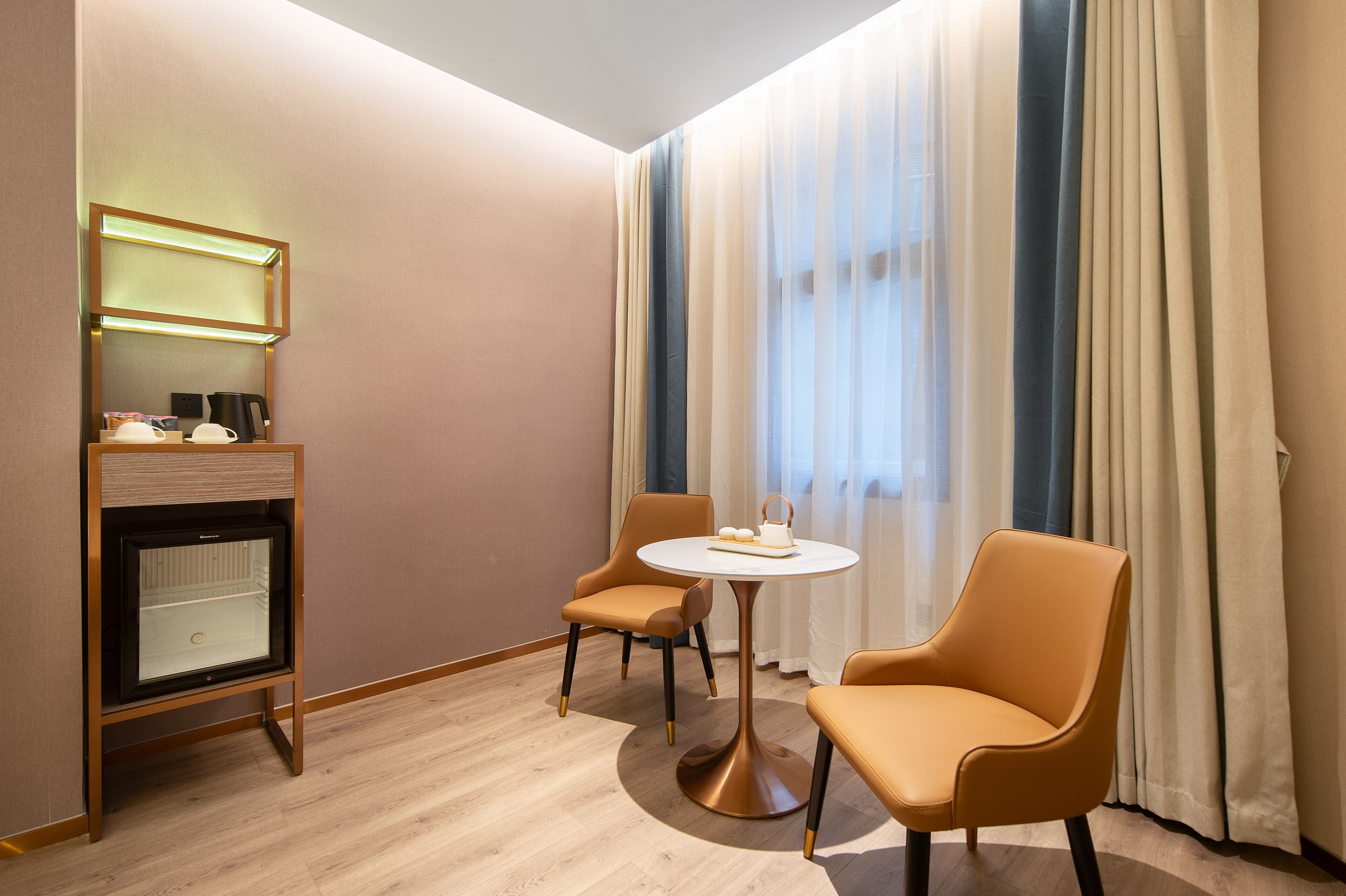A hotel room with a wooden floor, a round table with two chairs, a small cabinet with a mirror, and a window with sheer curtains.