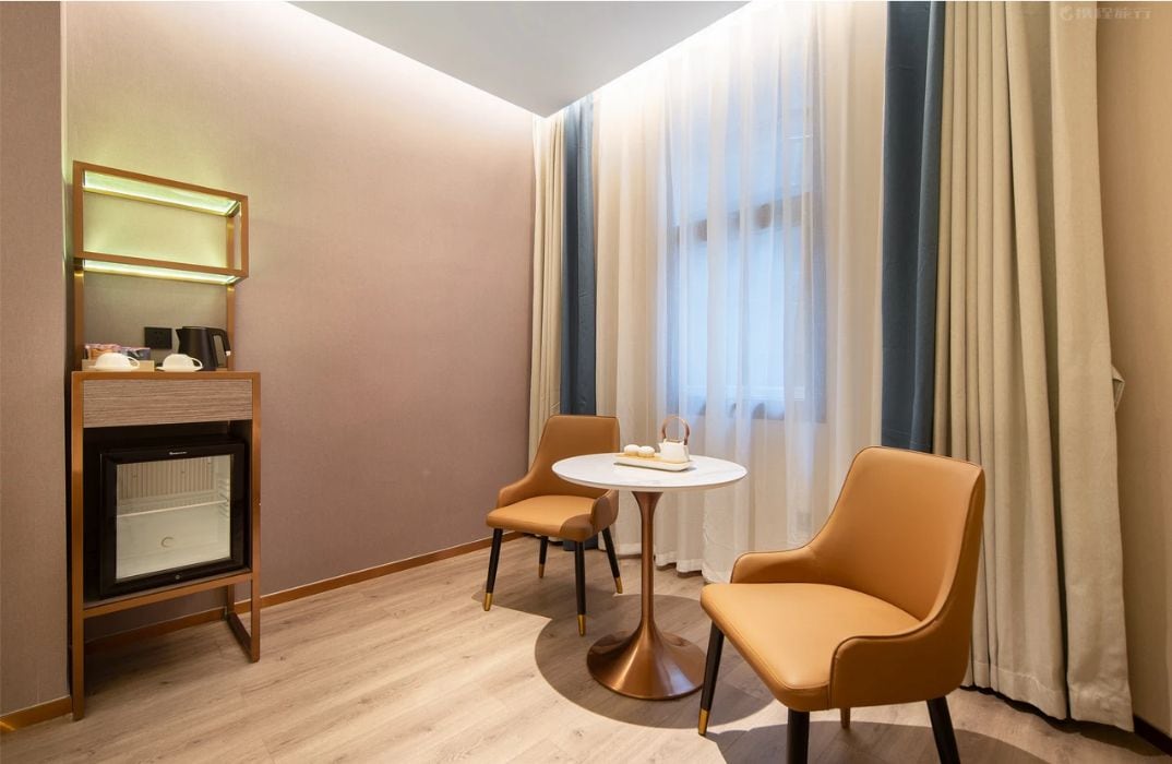 A hotel room with a minibar, a round table with two chairs, and a large window with sheer curtains.