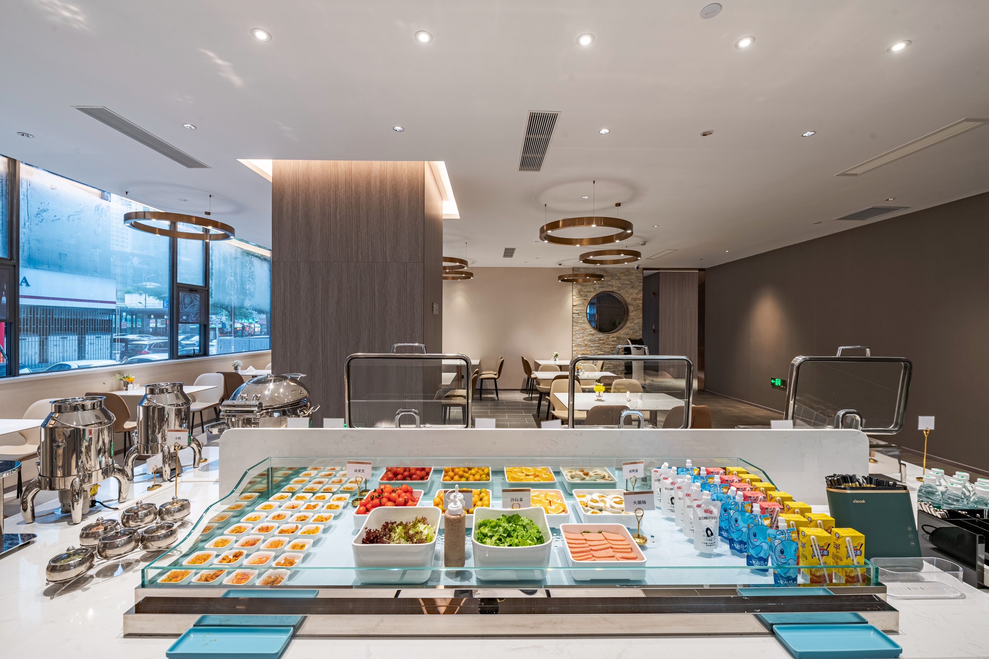 A modern hotel dining area with a buffet setup, including various food items and beverages, and a seating area with tables and chairs.