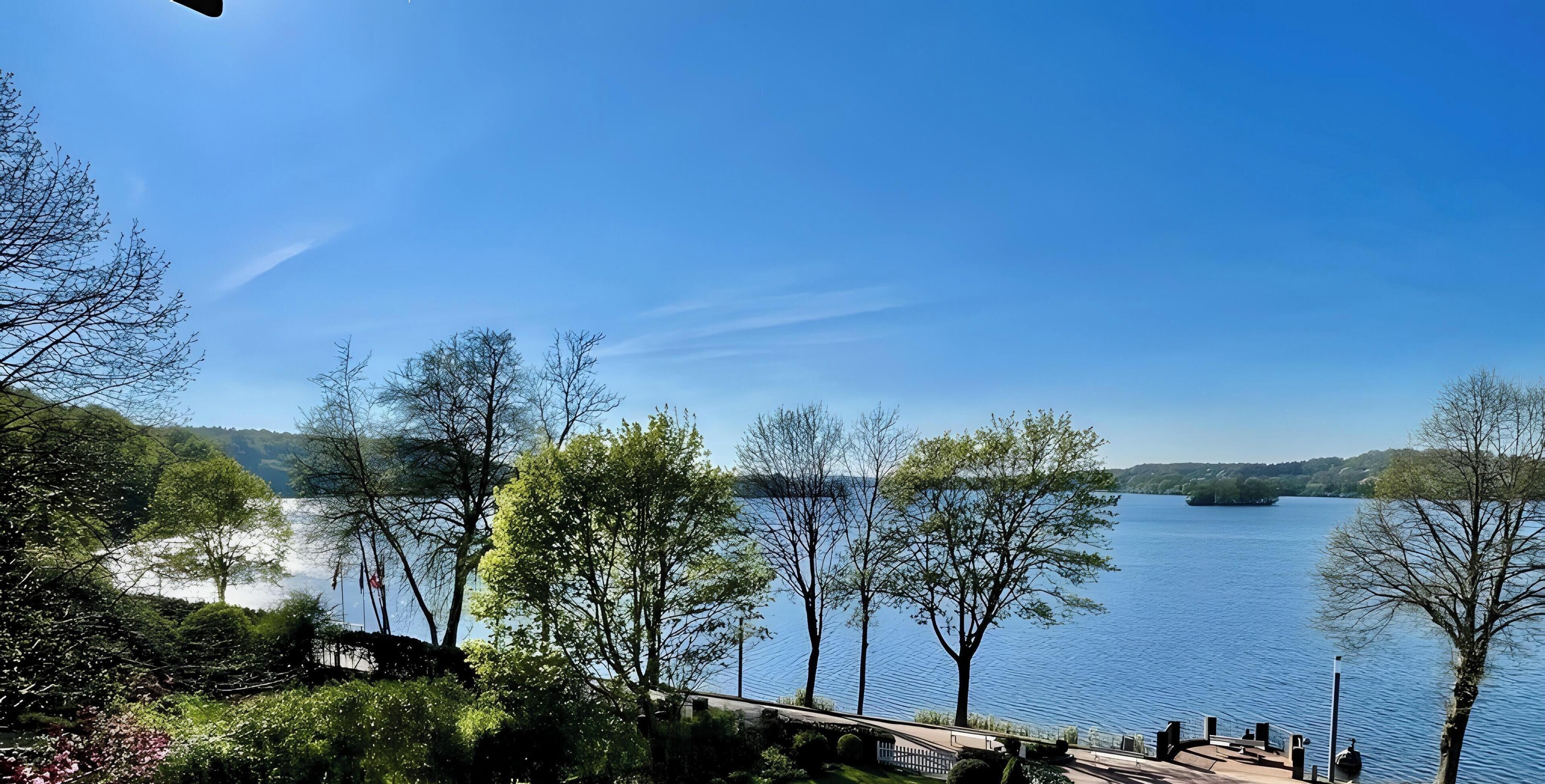 Uitzicht op een meer met een houten terras, bomen en een heldere lucht.