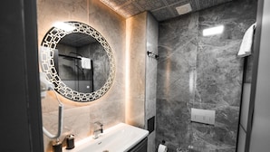 Un baño moderno con lavamanos de mármol, un espejo redondo con marco geométrico, ducha con panel de control y un inodoro.