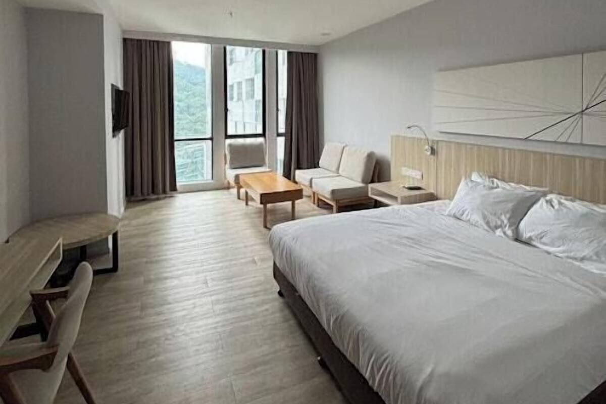 A modern hotel room with a large bed, a desk, a sofa, and a view of the outdoors.