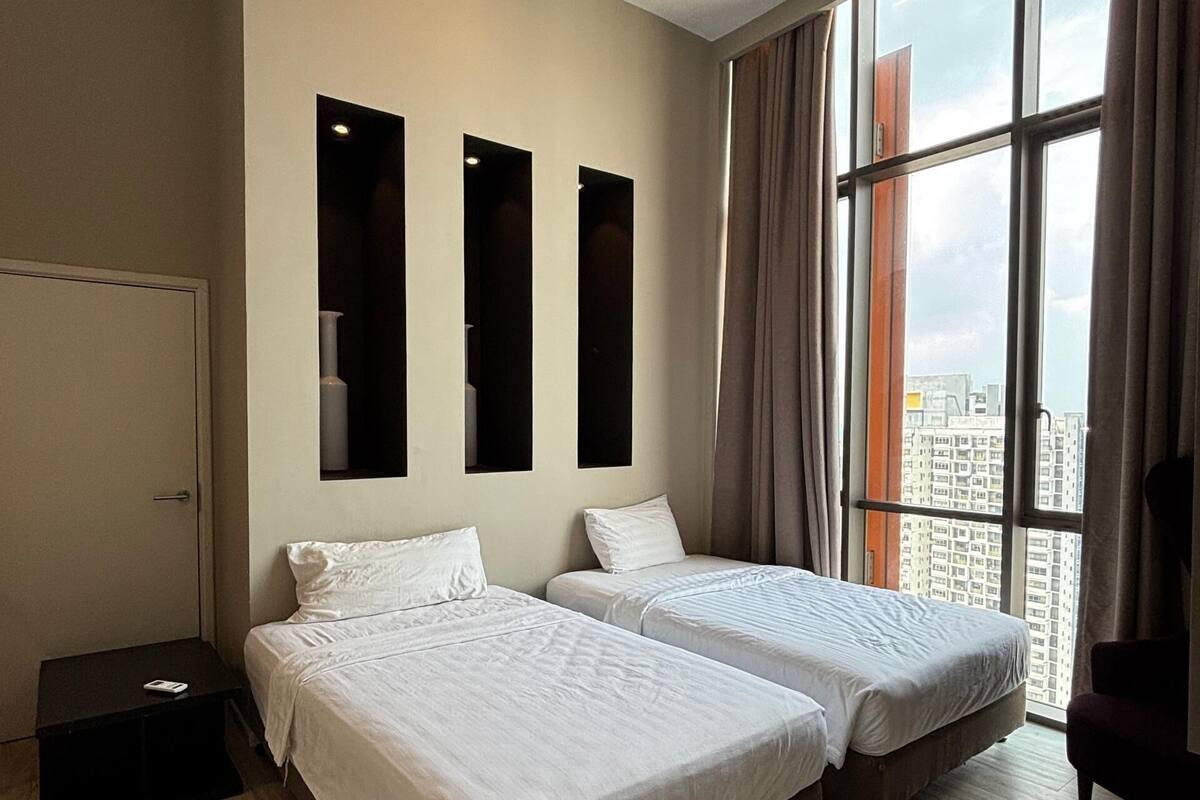 A hotel room with two beds, a small table, a chair, and a large window with curtains.
