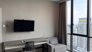 A modern hotel room with a large window, a flat-screen TV mounted on the wall, a grey sofa, a small desk with a chair, and a city view. - QQ Hotel Rooms Damansara Perdana (Petaling Jaya)