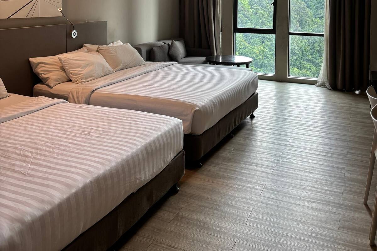 A modern hotel room with two beds, a sofa, a small table, and large windows offering a view of the forest.