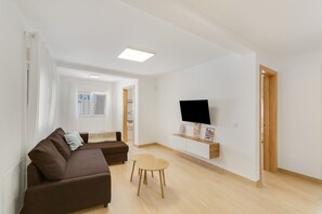 A modern living room with a brown sofa, a small wooden table, a flat-screen TV mounted on a white wall, and a wooden door.