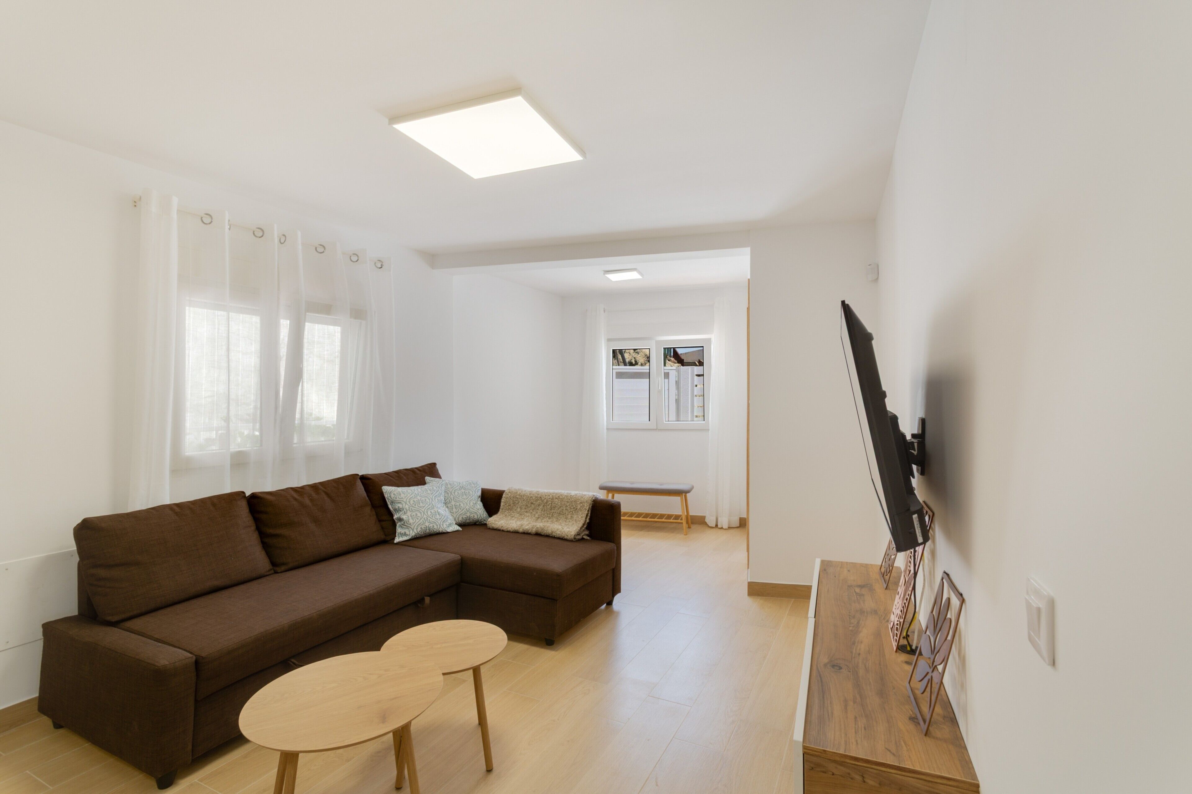A living room with a brown sectional sofa, a small wooden table, and a TV mounted on the wall.