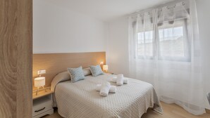 Una camera da letto con una testiera in legno, un letto con copriletto color beige, due abat-jour e una finestra con tende leggere.