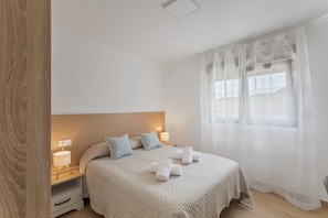 A bedroom with a wooden headboard, a bed with a beige quilt, two bedside lamps, and a window with sheer curtains.