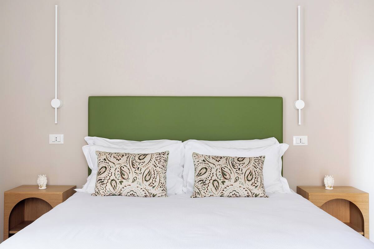 A neatly made bed with white linens, a green headboard, and patterned pillows. There are two bedside tables with decorative items and a wall-mounted light fixture.