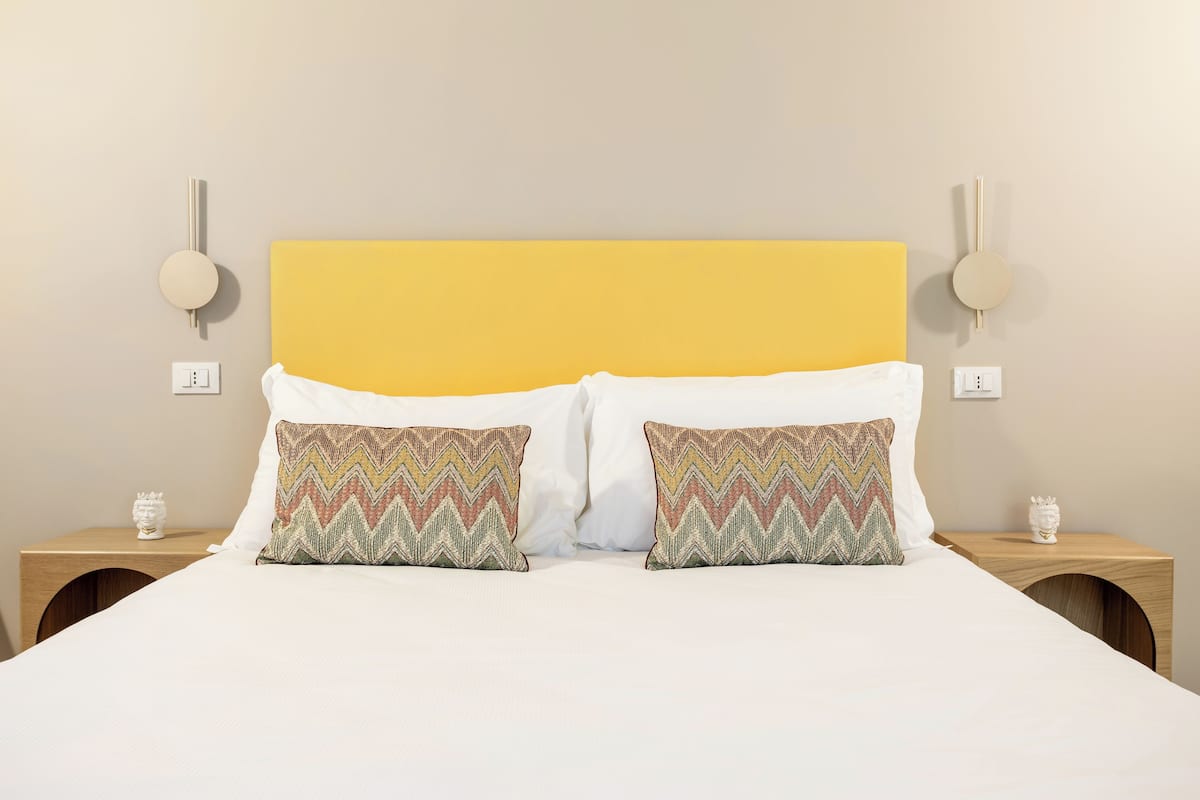 A bed with a yellow headboard, two patterned pillows, and wooden nightstands with wall-mounted lamps.