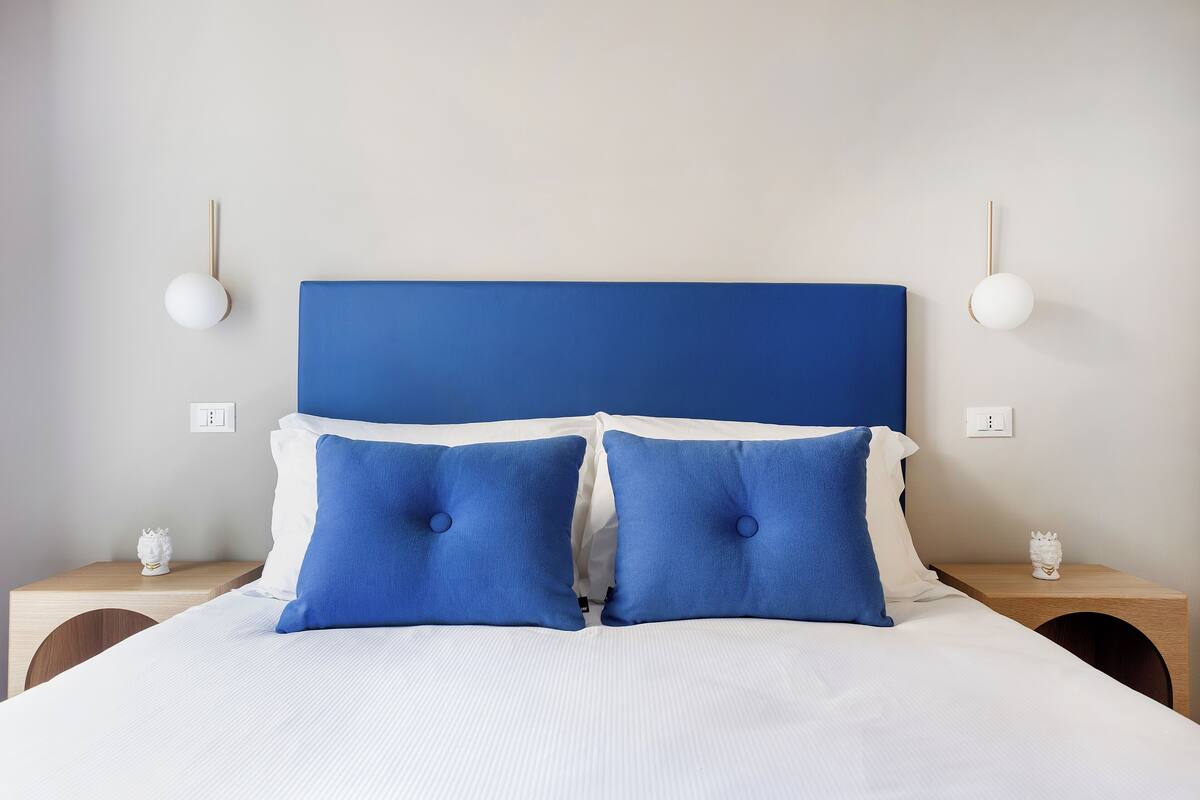 A neatly made bed with a blue headboard, two blue pillows, and a white comforter. There are two bedside tables with white lamps and a small decorative item on each.
