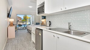 A modern kitchen with white cabinetry, a stainless steel sink, and a microwave. There is a dining area with a table and chairs, and a view of the outdoors through a glass door. - Pacific Blue Unit 175 Dual Swim Up B (Salamander Bay)
