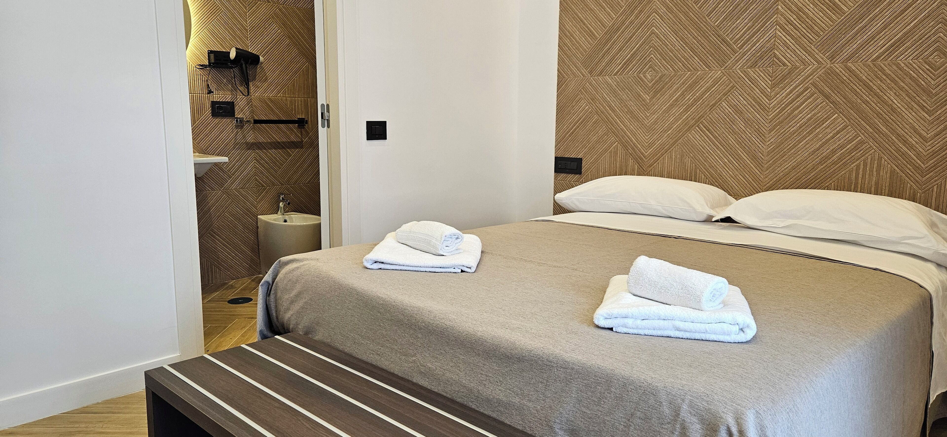 Hotelkamer met een bed, twee kussens, een nachtkastje, een bankje, een muur met een geometrisch patroon, een wastafel en een spiegel.