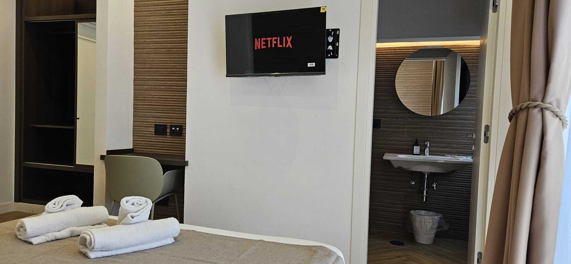 A hotel room with a bed, two rolled towels, a chair, a wall-mounted Netflix sign, a sink, and a trash bin.