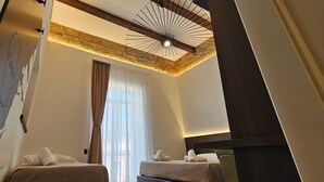 Habitació d'hotel amb dos llits, un mur de pedra decoratiu, un ventilador de sostre i una finestra gran amb cortines.