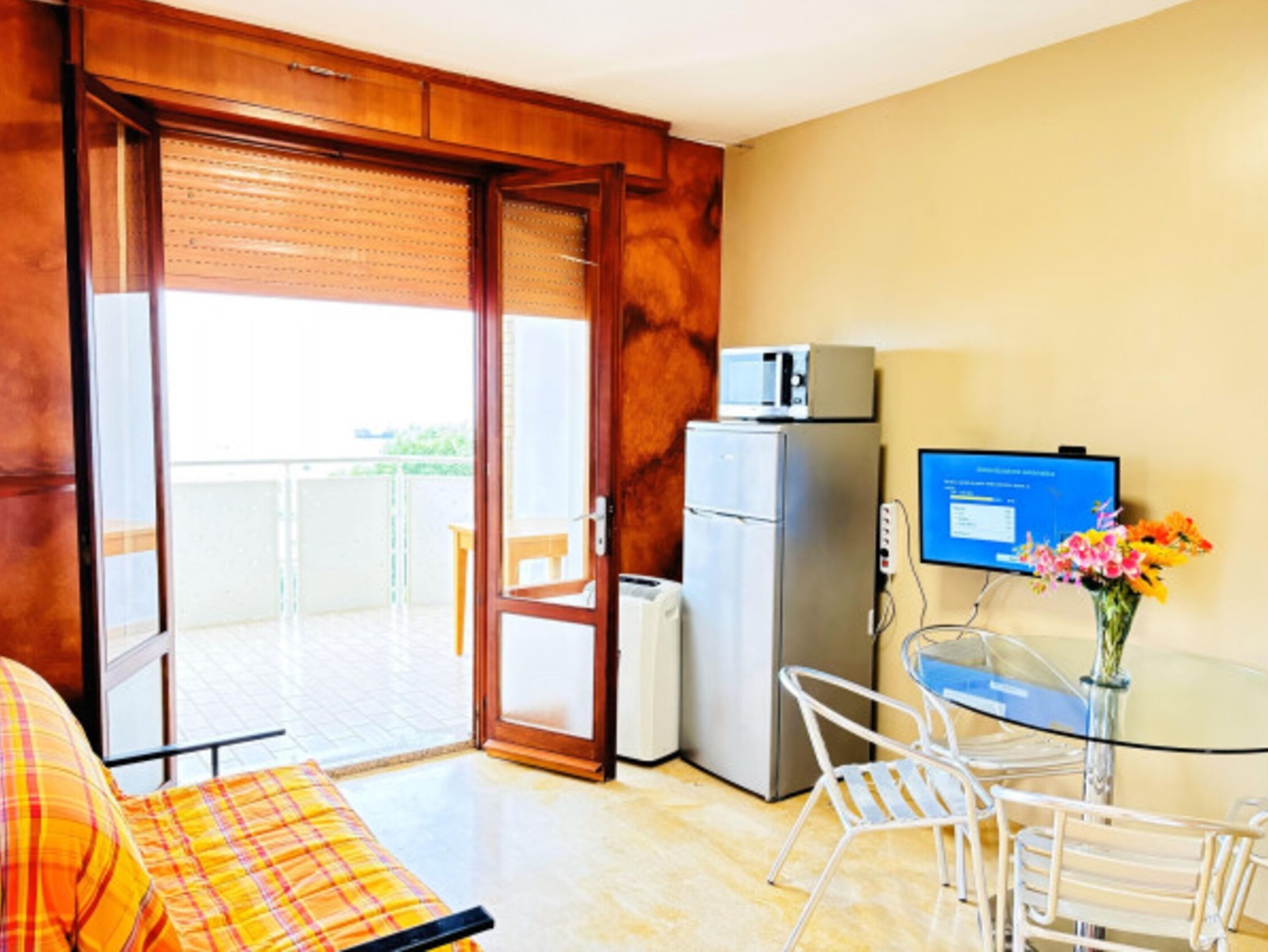 Ein Zimmer mit einem Bett, einem Schreibtisch mit Computer, einem Kühlschrank und einer Glastür zum Balkon.