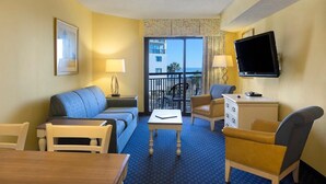 Une chambre d’hôtel avec un tapis bleu, un canapé, un fauteuil, une petite table, une télévision et un balcon offrant une vue sur les immeubles et les palmiers.