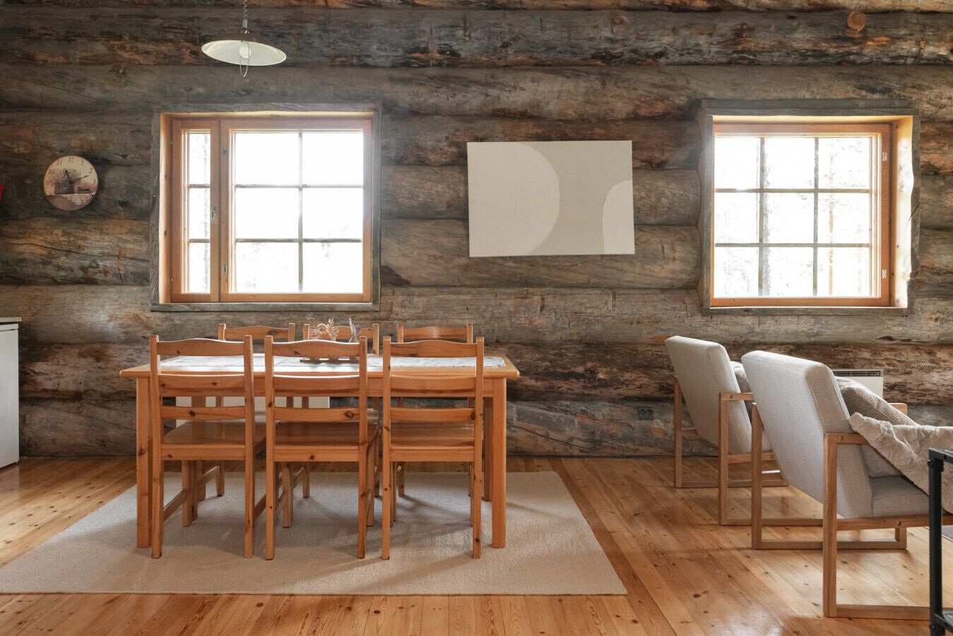 Ein rustikales Blockhaus-Interieur mit einem Holzespeisesaal und Stühlen, zwei Fenstern mit weißen Rahmen und einer Uhr an der Wand.