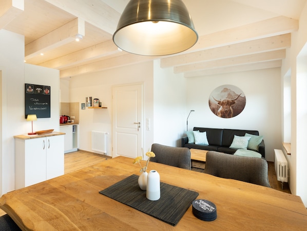 A modern living space with a wooden dining table, a sofa, a kitchen area, and a wall-mounted artwork.