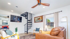 Un salon moderne avec un canapé en cuir marron, un fauteuil blanc, une table basse en bois, un tapis à motifs, un escalier en colimaçon noir et un ventilateur de plafond.