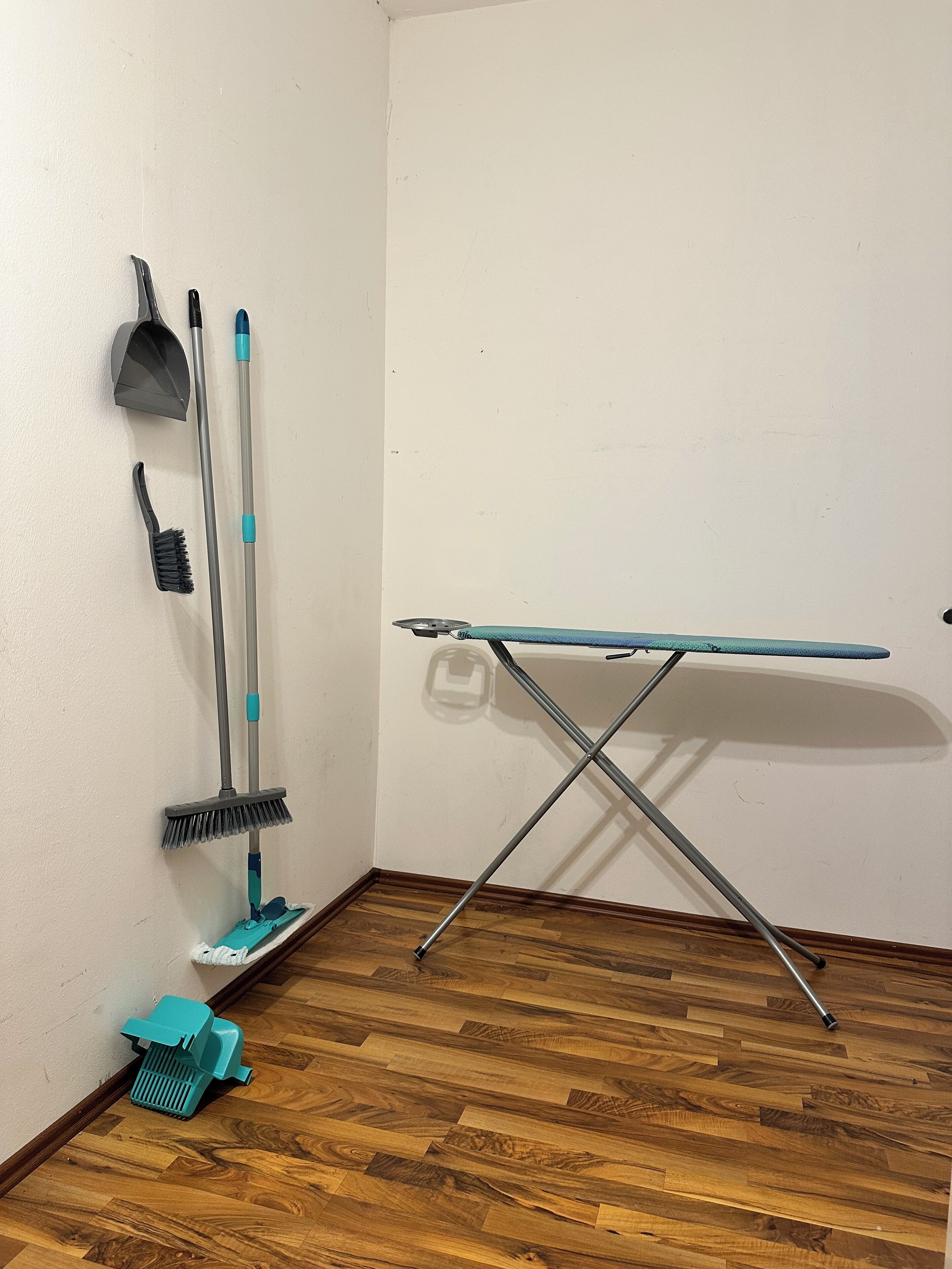 A corner of a room with a wooden floor, a wall-mounted ironing board, and cleaning tools including a broom, dustpan, and mop.