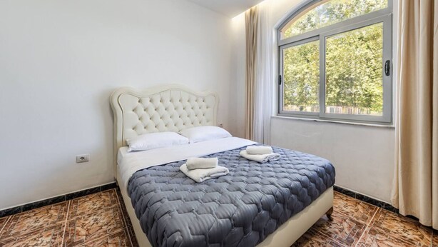 A neatly made bed with a tufted headboard and a quilted bedspread, a window with curtains, and a patterned floor. - Shiroka Stays 3 in Shkod r (Shkodër)
