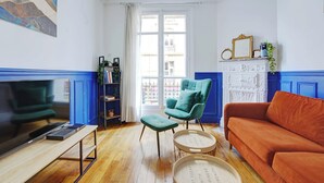 A living room with a television, a teal armchair, an orange sofa, and a white cabinet with a mirror and plant. - Elegant Apartment 1br/4p - Paris 15/montparnasse (Paris)