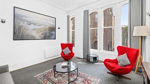 A modern living room with a large landscape painting, two red armchairs, a glass coffee table, and a patterned rug. - Steeple View Apartment (Montrose)