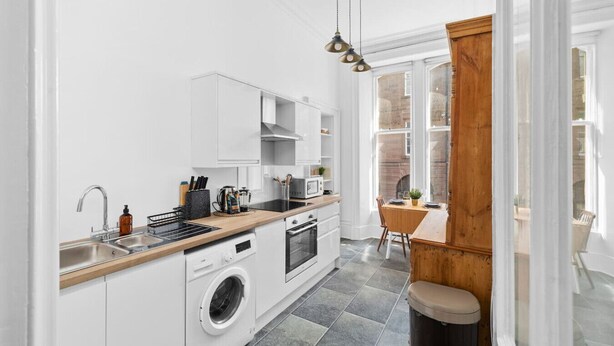 A modern kitchen with white cabinetry, a built-in washing machine, and a dining area with a wooden table and chairs. - Steeple View Apartment (Montrose)