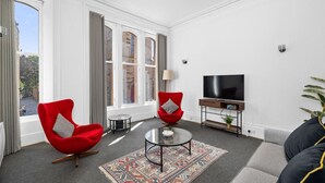 A modern living room with two red armchairs, a glass coffee table, a flat-screen TV, and a large window with curtains. - Steeple View Apartment (Montrose)