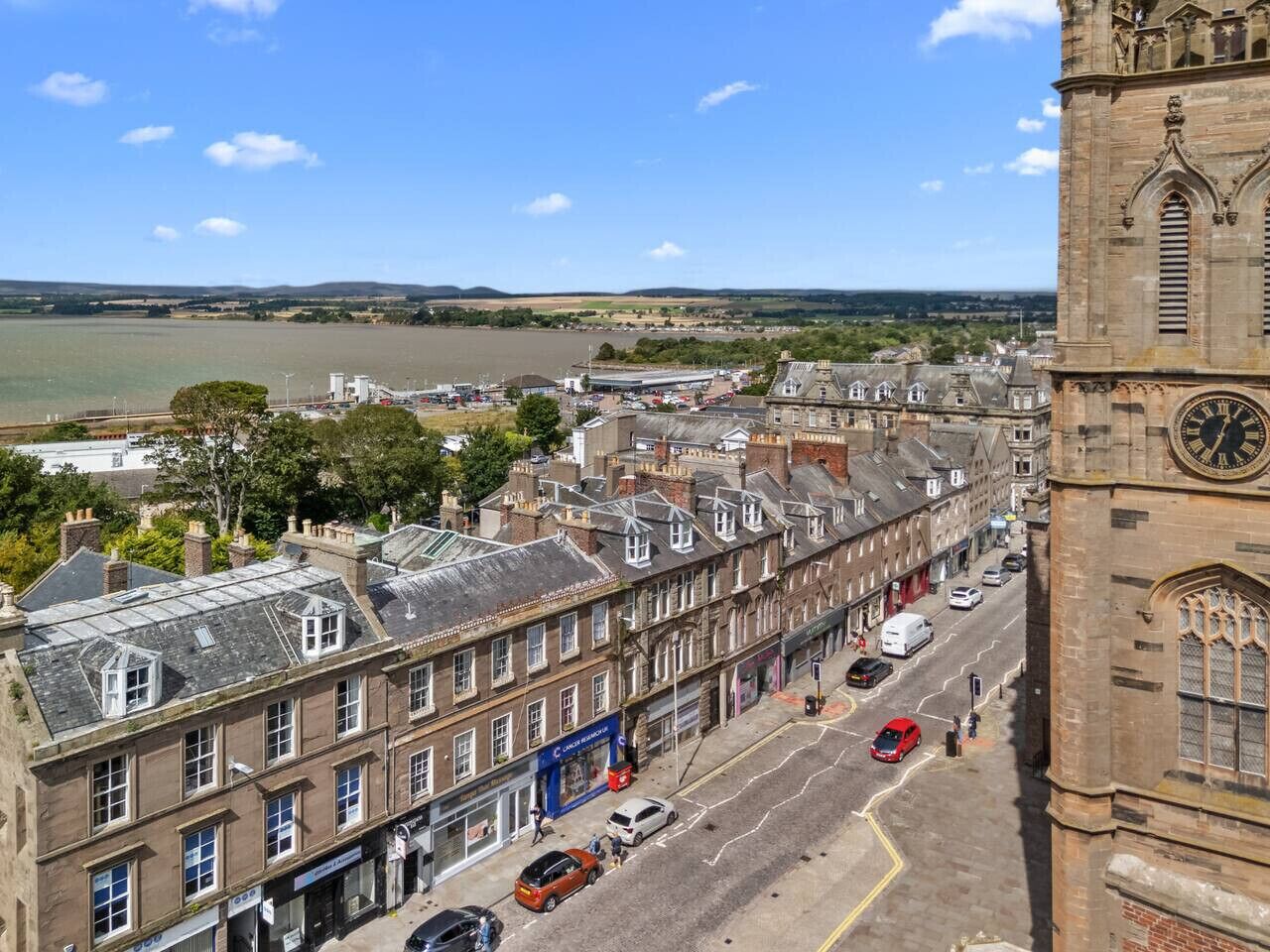 A historic building with a clock tower, a river in the background, and a street with cars and shops.
