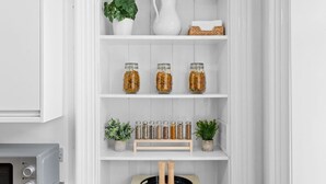 A white kitchen cabinet with shelves containing jars, a potted plant, a white pitcher, and a woven basket. - Steeple View Apartment (Montrose)