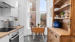 A kitchen with white cabinets, a wooden dining table, a blue bread bin, and a view of a brick building outside. - Steeple View Apartment (Montrose)