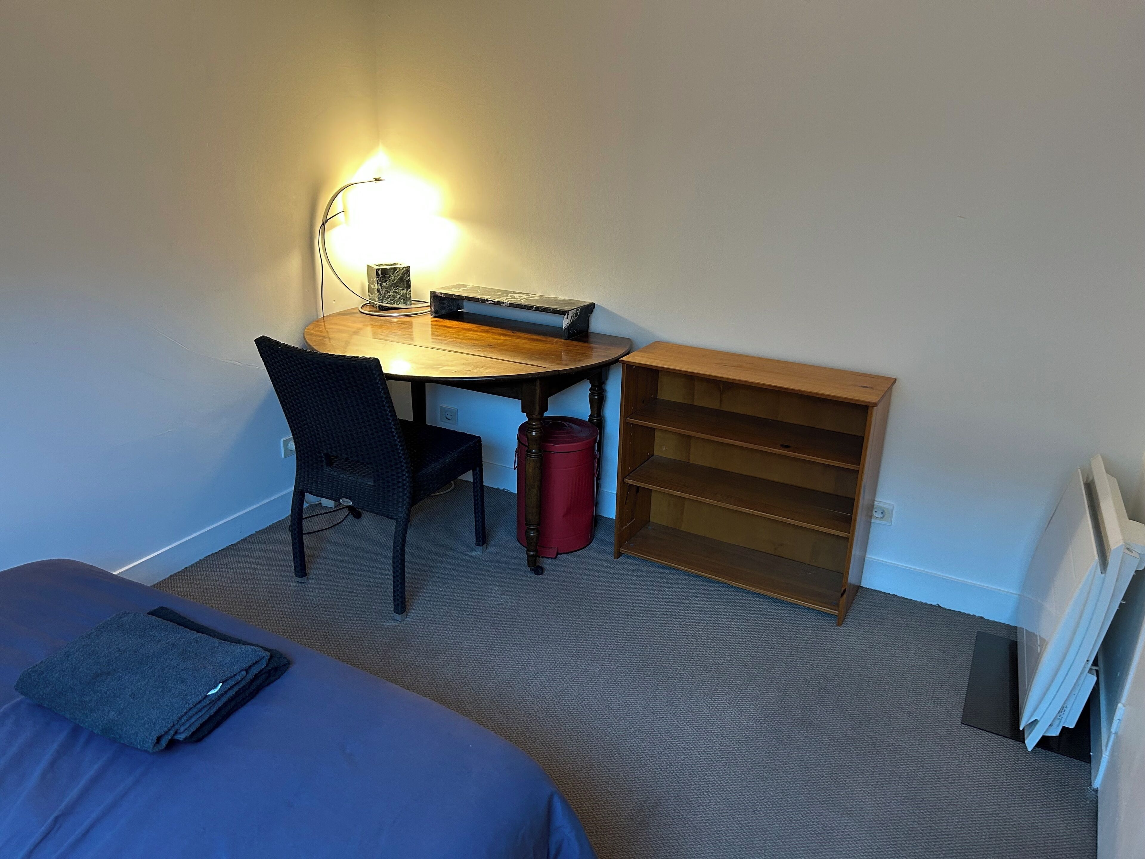Une chambre d’hôtel comprenant un lit, un bureau avec une chaise, une petite table avec une lampe, une poubelle rouge et une étagère en bois.
