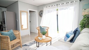 A living room with a wicker sofa, a round wooden coffee table, and a large window with sheer curtains. - Cosy Private Cottages- 3 bedrooms, pool and dstv  (Durban North)