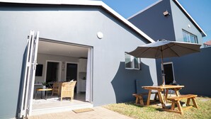 A modern house with a patio area featuring a wooden table and chairs, a large umbrella, and a sliding glass door leading to an interior space with a kitchen and dining area. - Cosy Private Cottages- 3 bedrooms, pool and dstv  (Durban North)