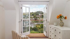 A room with a large open door leading to a balcony with a view of houses and trees. There is a chair and a dresser with drawers.