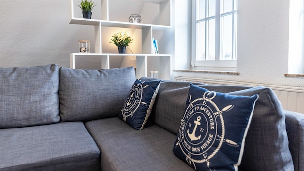 Een grijze bank met blauwe kussens, een wit wandrek met decoratieve objecten, en een raam met jaloezieën.