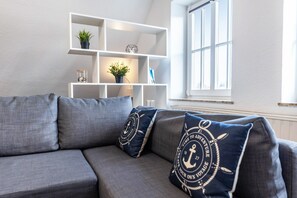 A gray sofa with navy blue pillows, a white shelving unit with decorative items, and a window with blinds. - Apartment 'Louis Hafenblick' with Sea View (Eckernförde)